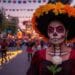 A woman in elegant Day of the Dead Catrina makeup and a floral hat, holding a white rose, with a parade